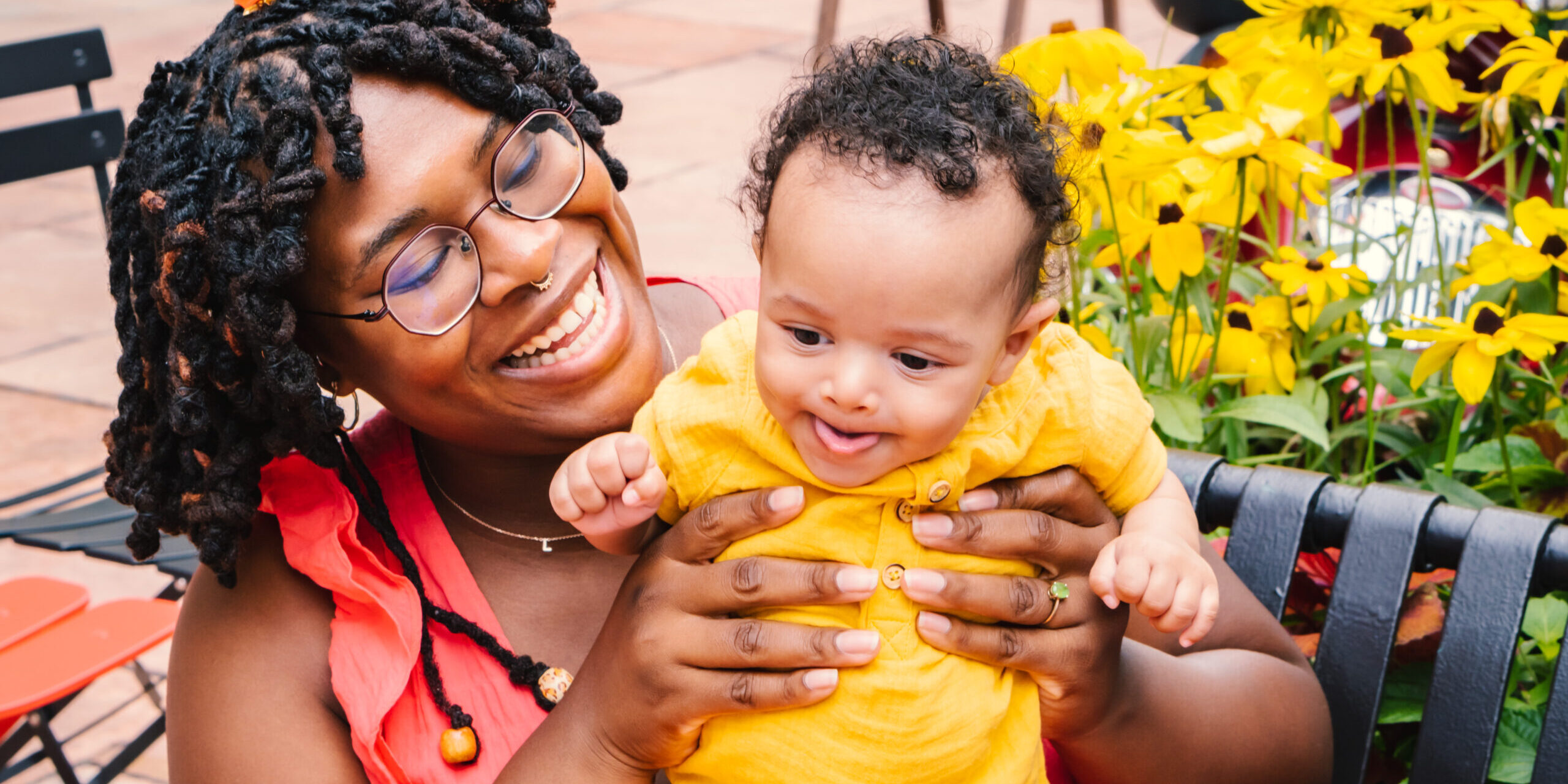 Black mother holding her baby wearing bright pink and yellow clothes sitting on a bench with flowers behind it.