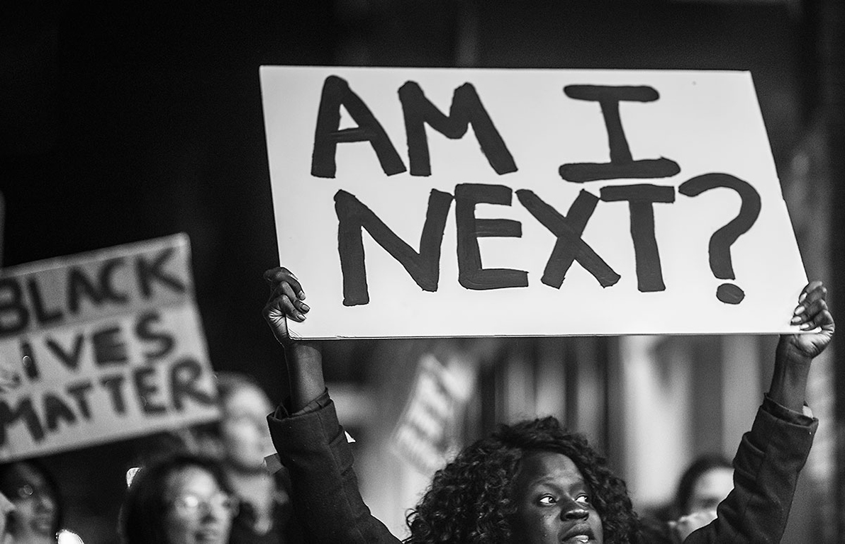 Black woman holding a sign that says "Am I Next?"