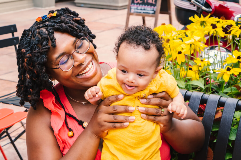 Black mother and her baby wearing bright yellow and pink clothing sitting on a bench